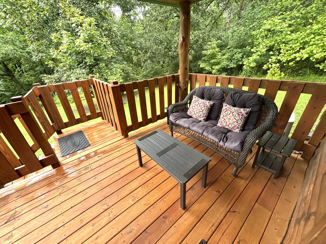 A cushioned wicker loveseat and low table on the deck at Red Oak Cabin, woods beyond the rail