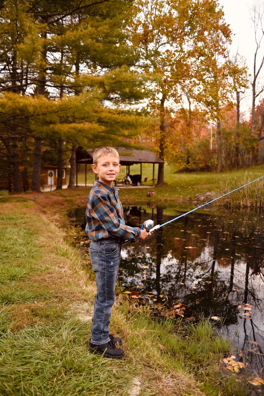 A young guest fishing from the bank of Red Oak Cabin's own stocked pond, with the picnic shelter and autumn woods behind
